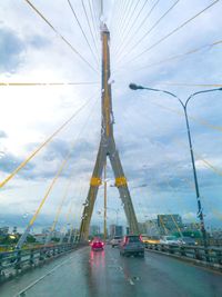 Cars on suspension bridge over road against sky