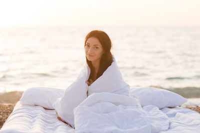 Smiling woman sitting in bed with duvet and pillow over sea background outdoors. 