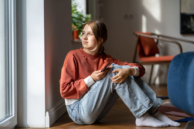 Pensive girl teenager sits on floor looks at window with smartphone in hand at home.