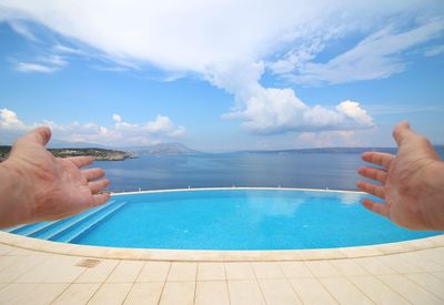 Cropped hands reaching swimming pool and sea against sky