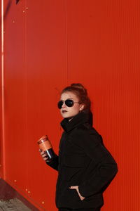 Fashionable teenage girl wearing black warm clothing standing against red wall during sunny day