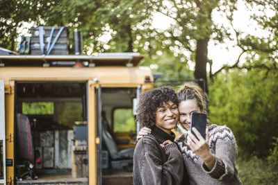 Woman taking selfie with friend against caravan during camping in forest