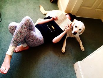 High angle view of woman holding book in front of face while relaxing on dog at home