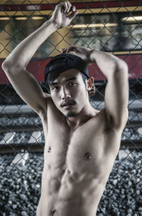 Portrait of shirtless man leaning on chainlink fence against train