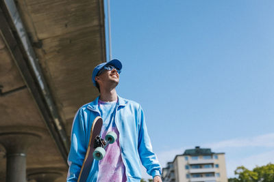 Low angle view of fashionable man in casuals looking up while standing under blue sky near bridge