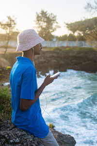 Side view of young man standing by ocean using a mobile phone