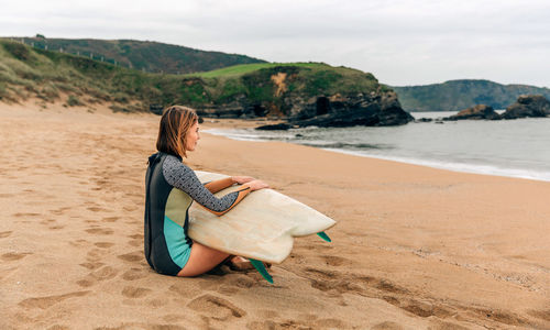 Surfer woman with wetsuit and surfboard sitting on the sand looking at the sea