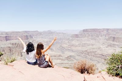 Rear view of women with arms raised sitting on landscape during sunny day