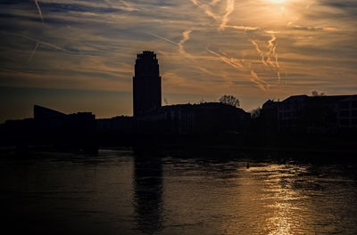 Silhouette buildings by river against sky during sunset