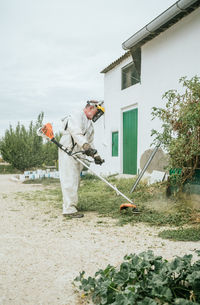 Full body side view of aged unrecognizable male farmer in white coverall clothes and protective mask cutting grass with weed cutter while working in garden on cloudy summer day