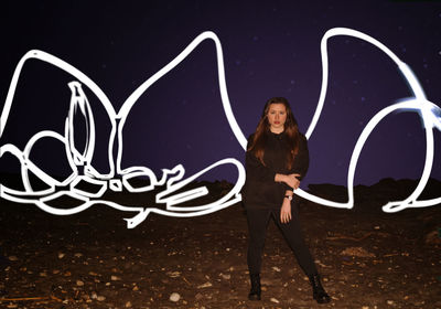 Portrait of young woman with light painting standing on field against sky at night