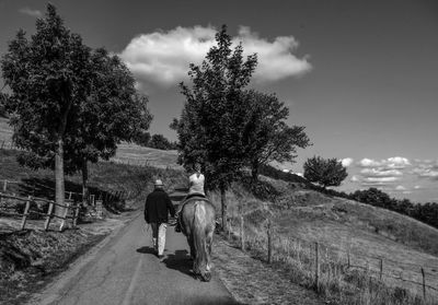 Girl riding horse with man on road amidst trees against sky