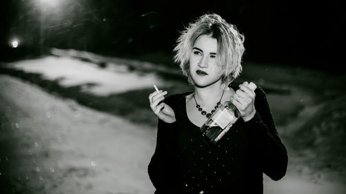 Close-up of young woman smoking cigarette while holding alcohol bottle at night