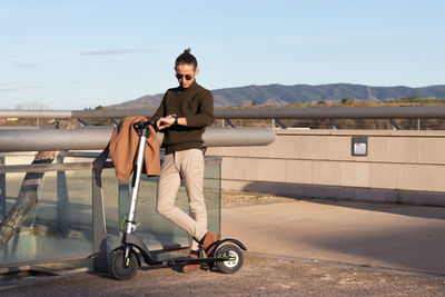 Young man using smartwatch and with electric scooter in the city park with mountains behind