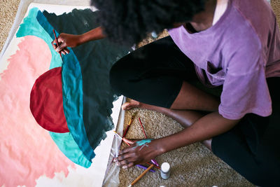 From above of crop anonymous black female with paintbrush drawing colorful picture on paper while sitting on floor at home