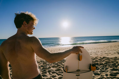 Rear view of shirtless man with surfboard at beach during sunset