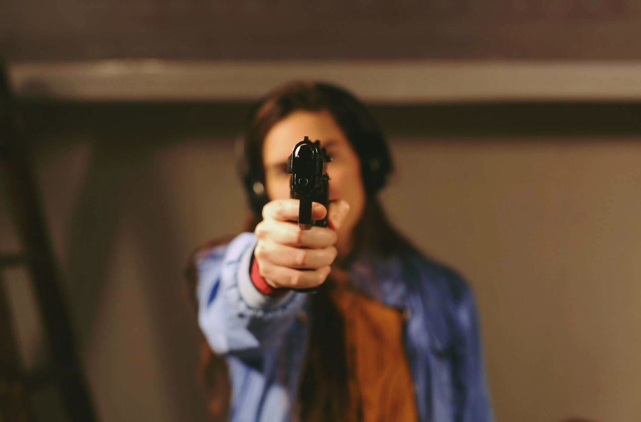 one person, holding, front view, gun, weapon, aiming, waist up, portrait, standing, indoors, young adult, real people, handgun, adult, casual clothing, communication, technology, activity, focus on foreground, digital camera, hairstyle, aggression