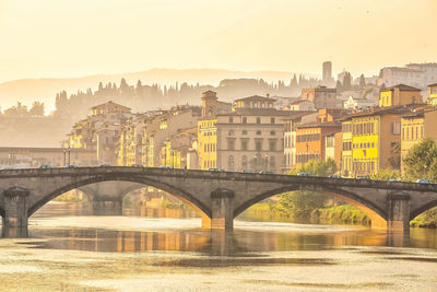 Bridge over river by buildings against sky during sunset