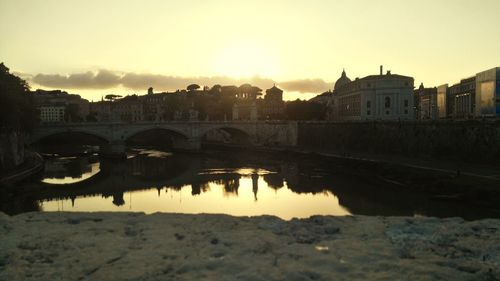 Bridge over river by buildings against sky during sunset