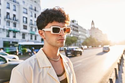 Young male in white shirt and trendy sunglasses looking away on blurred background of road illuminated with sunset sky in city