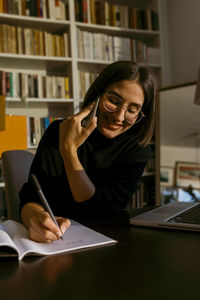 Smiling businesswoman talking on smart phone while writing in book sitting at home
