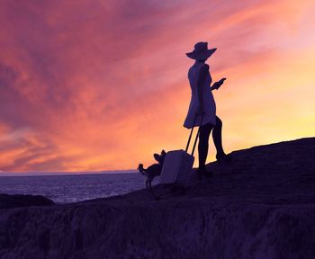 Woman holding suitcase by dog standing on cliff at beach against sky during sunset