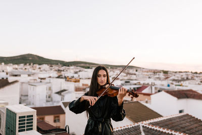 Young beautiful female musician in stylish mini dress holding acoustic violin and standing on rooftop in residential suburb and closed eyes on sunny evening