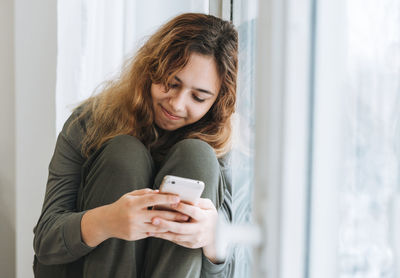 Beautiful smiling teenager girl with curly hair using mobile phone sitting on window sill, 