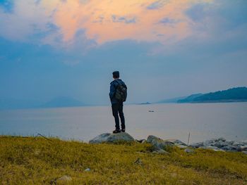 Rear view of man standing on rock by sea against sky at sunset