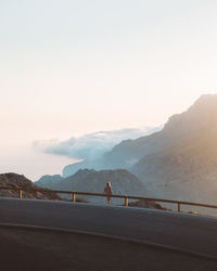 Rear view of woman sitting on railing on road by mountains against clear sky