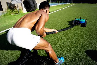 Rear view of shirtless athlete pulling rope during crossfit training on grass