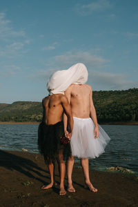 Shirtless gay couple wearing skirts while standing against lake