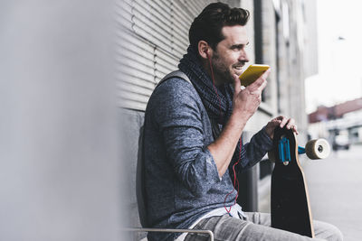 Businessman with skateboard sitting at bus stop using smartphone