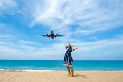 Woman standing below flying airplane at beach against sky