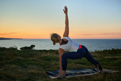 Rear view of woman exercising at beach against sky during sunset