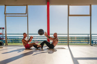 Side view of confident male athletes practicing with medicine ball and pumping abdomen muscles in spacious sports center