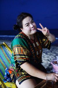 Portrait of woman showing peace sign while sitting on chair at beach during dusk