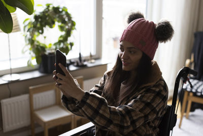 Woman wearing knit hat taking selfie through smart phone while sitting on wheelchair at home