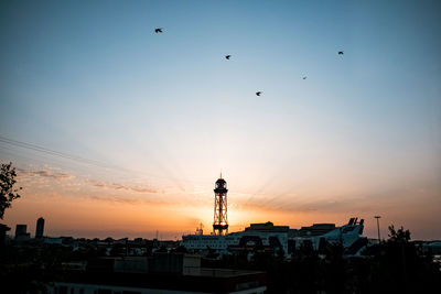 Silhouette birds flying by buildings against sky during sunset