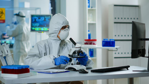 Scientist wearing mask sitting by microscope at laboratory