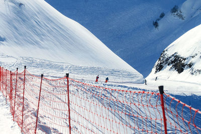 In the background of the orange grid fence is a gentle ski run with snowboarders and skiers.