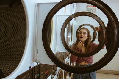 Young woman applying lipstick reflecting on washing machine