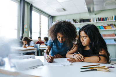 Curly hair girl writing while sitting with female classmate near desk in classroom at elementary school