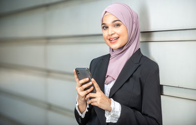 Portrait of smiling businesswoman using mobile phone while standing against wall