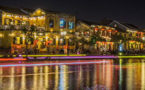 Illuminated light trails on street by buildings against sky at night