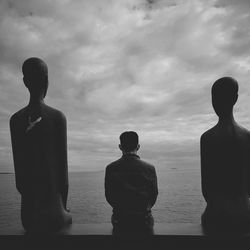 Man sitting amidst statues by sea against cloudy sky