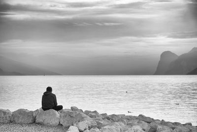 Rear view of man sitting on rock by sea against sky