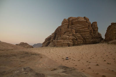 A 4x4 crosses the desert of wadi rum, between the immense rocks that surround it, against clear sky.