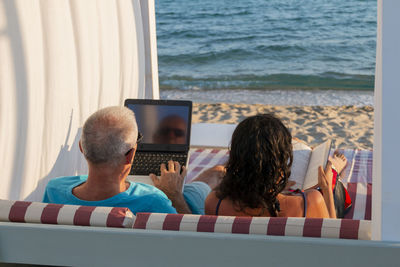 Man working on his laptop while his wife reading a book at the beach. business and summer vacation