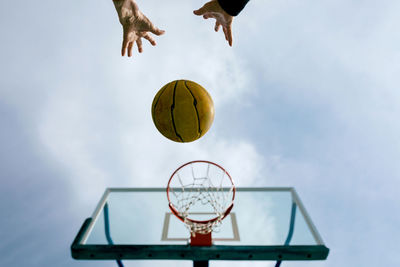 From below of crop anonymous person throwing basketball ball into hoop while playing game on public sports ground against blue sky